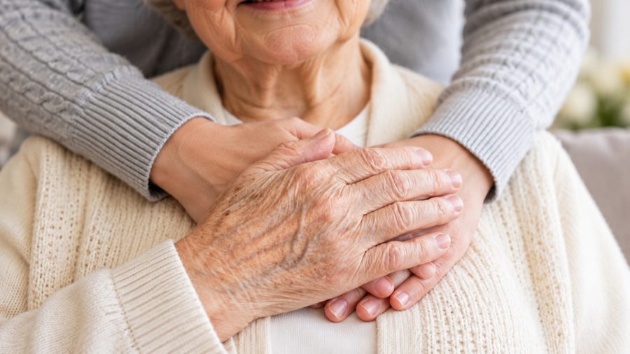 A younger person hugs an elderly woman from behind, their hands resting together over her heart; she is smiling, wearing a cream knit cardigan.