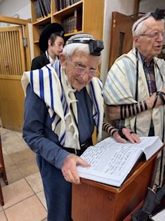 An elderly man wrapped in a tallis and wearing tefillin smiles as he reads from a large open prayer book on a stand, with other men praying behind him.