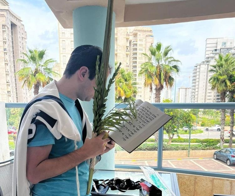 A young man wrapped in a tallis, holding a lulav and esrog, reads from a prayer book held close to his face on a sunny balcony with palm trees behind him.
